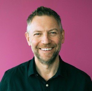 An image of nigel smiling infront of a pink background.