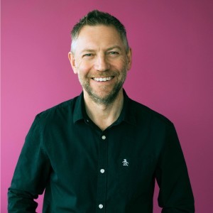 An image of nigel smiling infront of a pink background.