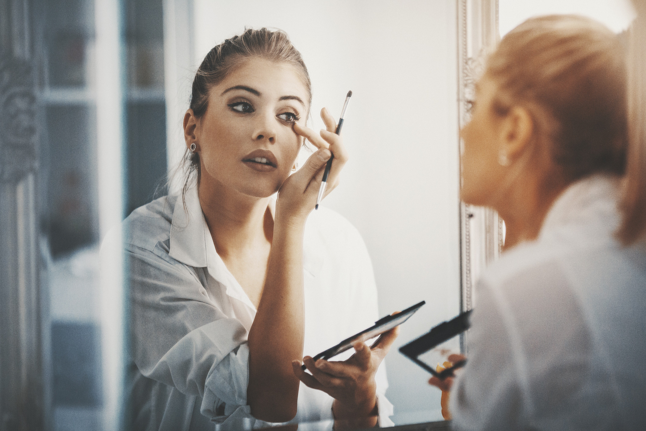 An image of a woman doing her makeup in the mirror after spring cleaning her makeup bag.