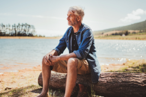 An image of a mature, older man sitting next to a lake doing nothing.