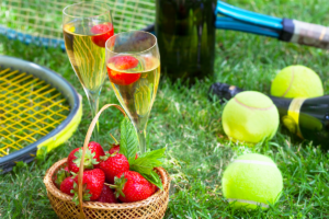 An image of a basket of strawberries next to two flutes of campaign and a tennis racket.