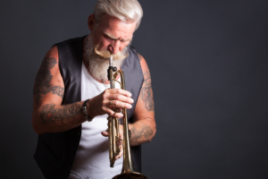 An image of a man over 50 with tattoos playing the trumpet.