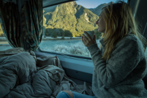 An image of a middle aged woman drinking a warm beverage in a camper van while looking out at the beautiful view.