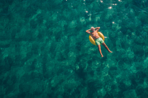 An aerial shot of a middle aged man relaxing on a yellow rubbber ring float in the deep blue ocean.