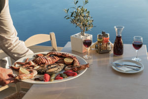 An image of a waiter serving a plate of seafood on a table with red wine, overlooking the Mediterranean sea.