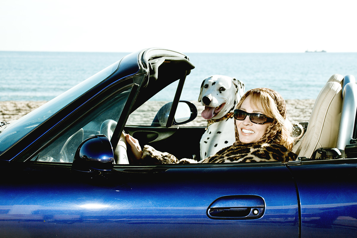 An image of a mature woman in a blue convertible with her dog sitting in the front.