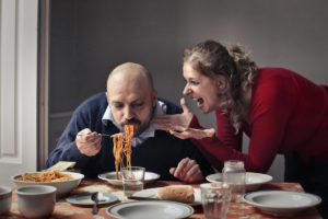 An image of a noisy man slurping spaghetti while his furious wife shouts at him.