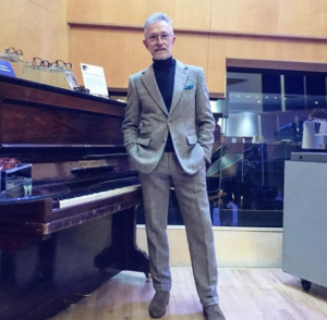 stylish mature man stood next to a piano in a high neck black top and fitted suit