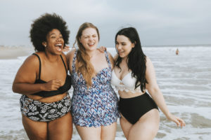 three cheerful plus size women laughing together on the shore of a beach