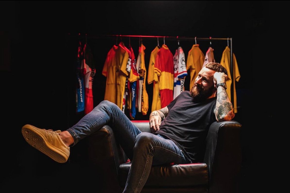 Bradley Wiggins sat in chair with rail of cycling clothes behind