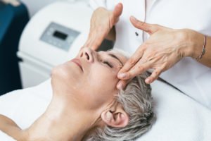 middle aged woman getting a facial treatment lay on bed with beautician massaging her head