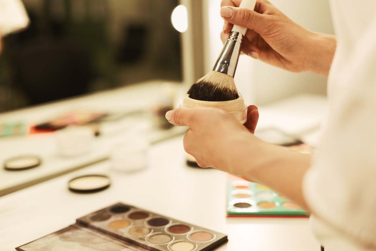 close up of woman holding a make up brush in powder with other make up products on the desk in front of her